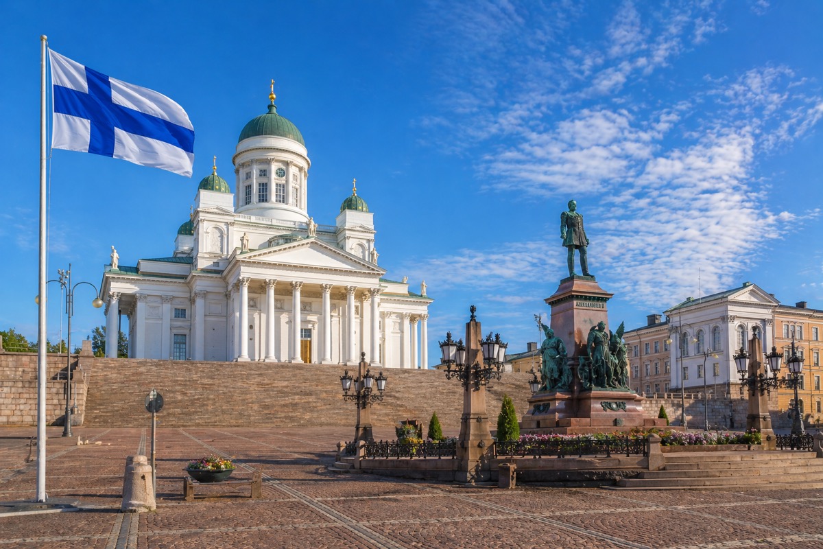 The Finnish flag waving in the foreground at Senate Square in Helsinki, Finland, featuring the iconic white Helsinki Cathedral and the Alexander II statue under a bright blue sky.